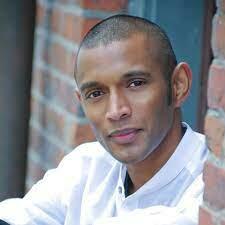 Leaning against a brick wall, wearing a lose white top, composer Dinuk Wijeratne looks into the camera.