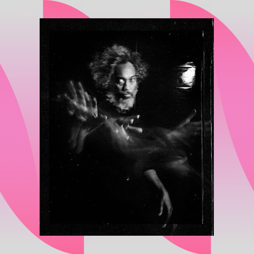 Image description: A black and white portrait photograph of multidisciplinary artist Zachary Fabri. Made with a large format camera as the grain of the film is visible. Fabri is a forty-some year old Black man with a graying afro and beard. His focused eyes glancing off to the side tell us that he is alert. His right hand swims through the space of the photo, captured in motion in a ghostly effect. The pattern of his hand movement is similar to that of a magician or music conductor. Fabri is stationed in a near entirely pitch black setting that is devoid of any visible objects or architecture. The only exception to this black void is a small light in the far distance of the image casting onto a metal, industrial surface. Fabri’s distance from this single light suggests an expansiveness to the space he is in.
