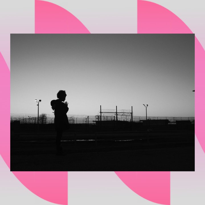 Image description: A black-and-white photograph that shows the silhouette of a person praying in front of a landscape of barbed wire fences, floodlights, and a guard tower on the far right.