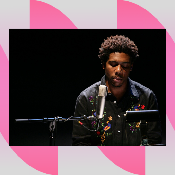 Image Description: A landscape-oriented photograph of a medium-dark complexioned black man sitting behind a microphone and a music stand with a lamp attached to the top. The man has a short afro and wears a black button-down shirt embroidered with brightly colored flowers and birds. The man sits in front of a black background.