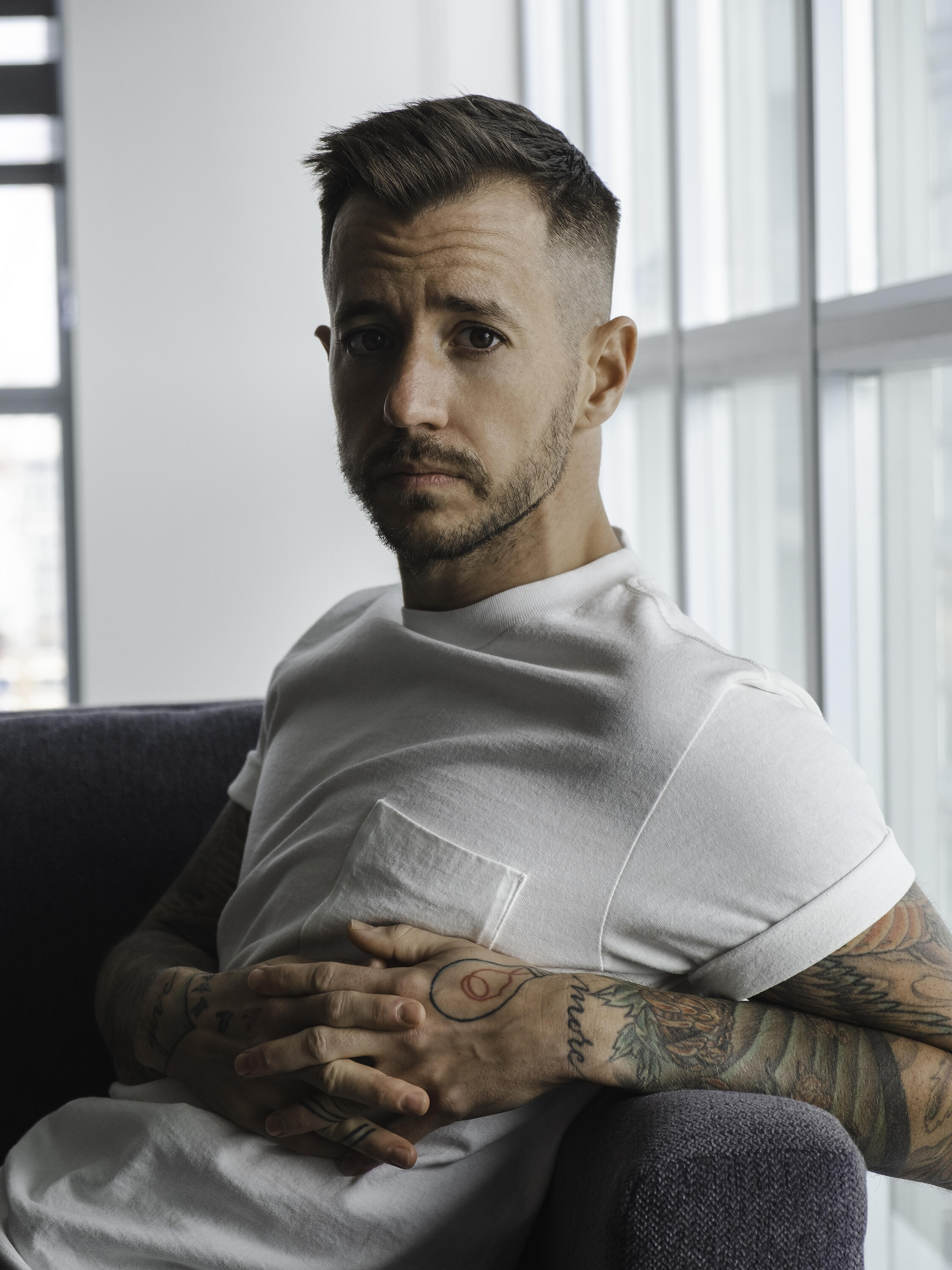 White man with dark hair looks at the camera. He is sitting in a well lighten room and has tattoos in his arms.