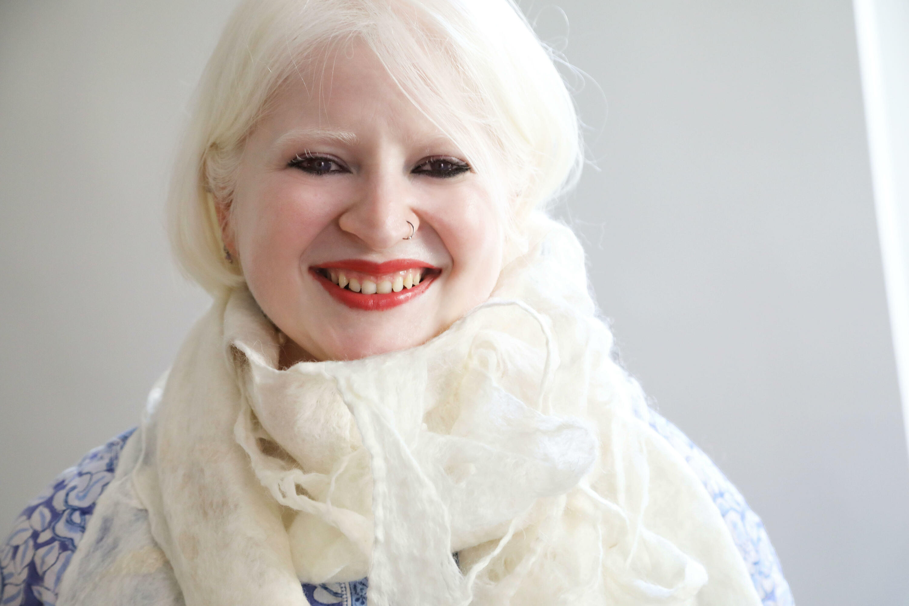 Image Description: Sugandha looks directly at the camera, smiling. She is wearing a white scarf, a white and blue patterned shirt, and red lipstick. 