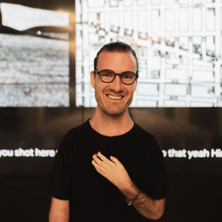 Josh is looking directly into the camera, smiling. They are wearing a black t-shirt and black framed glasses. They have their left hand crossed on their chest. Behind them is a black and white background.