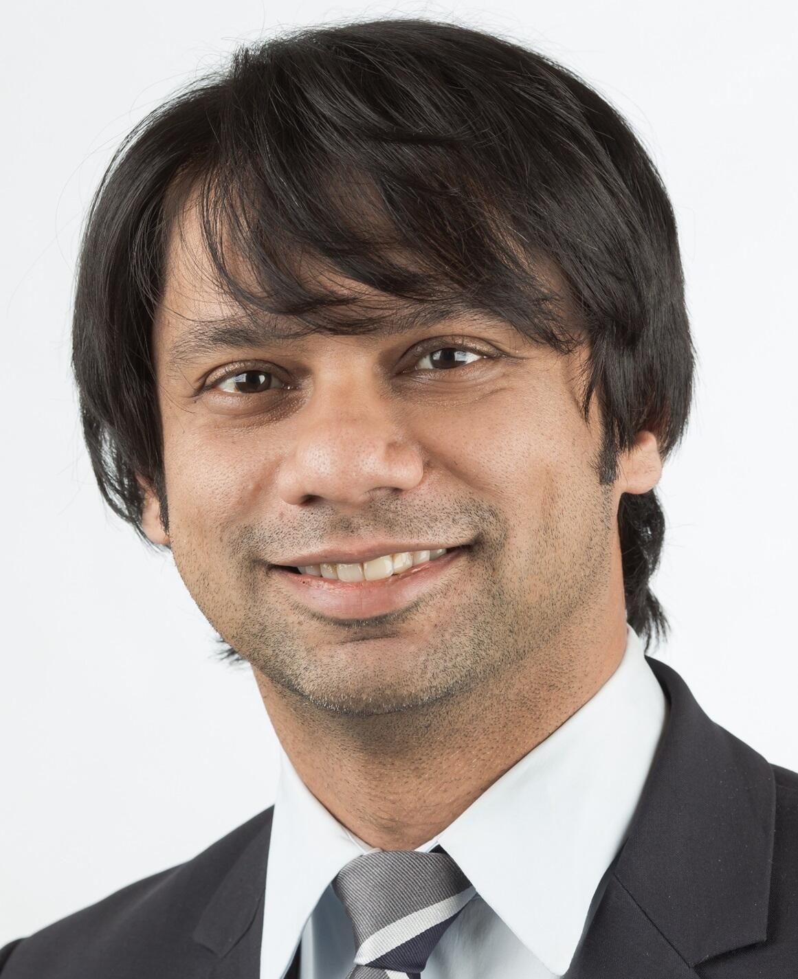 Gaurav is an indian man with dark black hair. He is wearing a suit.
