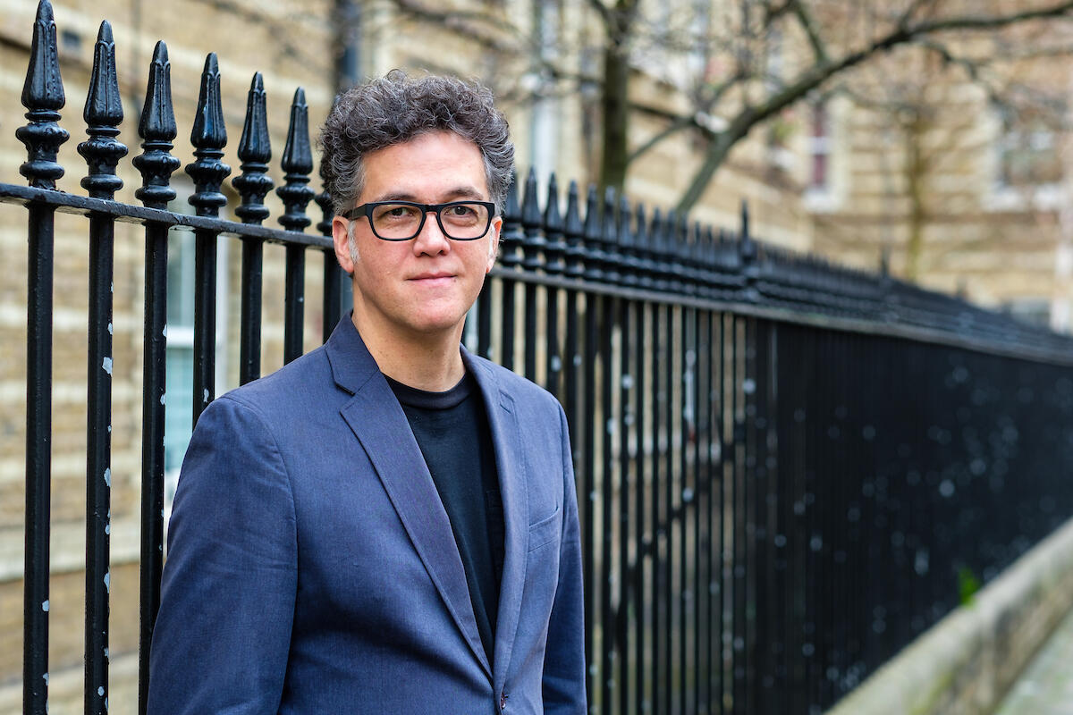 Carlos is a Caucasian man with dark hair and glasses. He is wearing a light blue suit coat and dark shirt. 