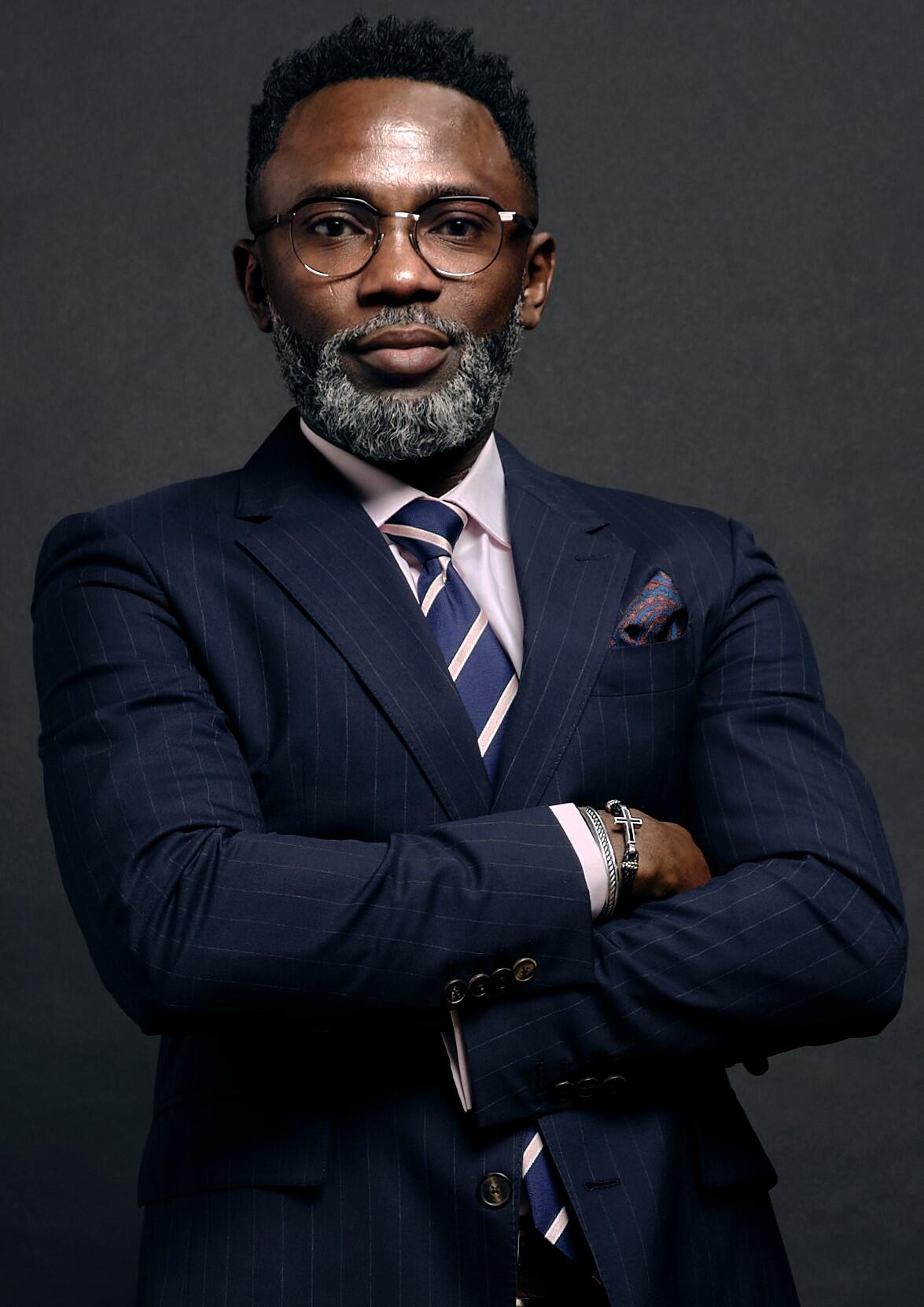 Lanre is a black man with dark hair, a beard, and glasses. He is wear a blue pin stripe suite.