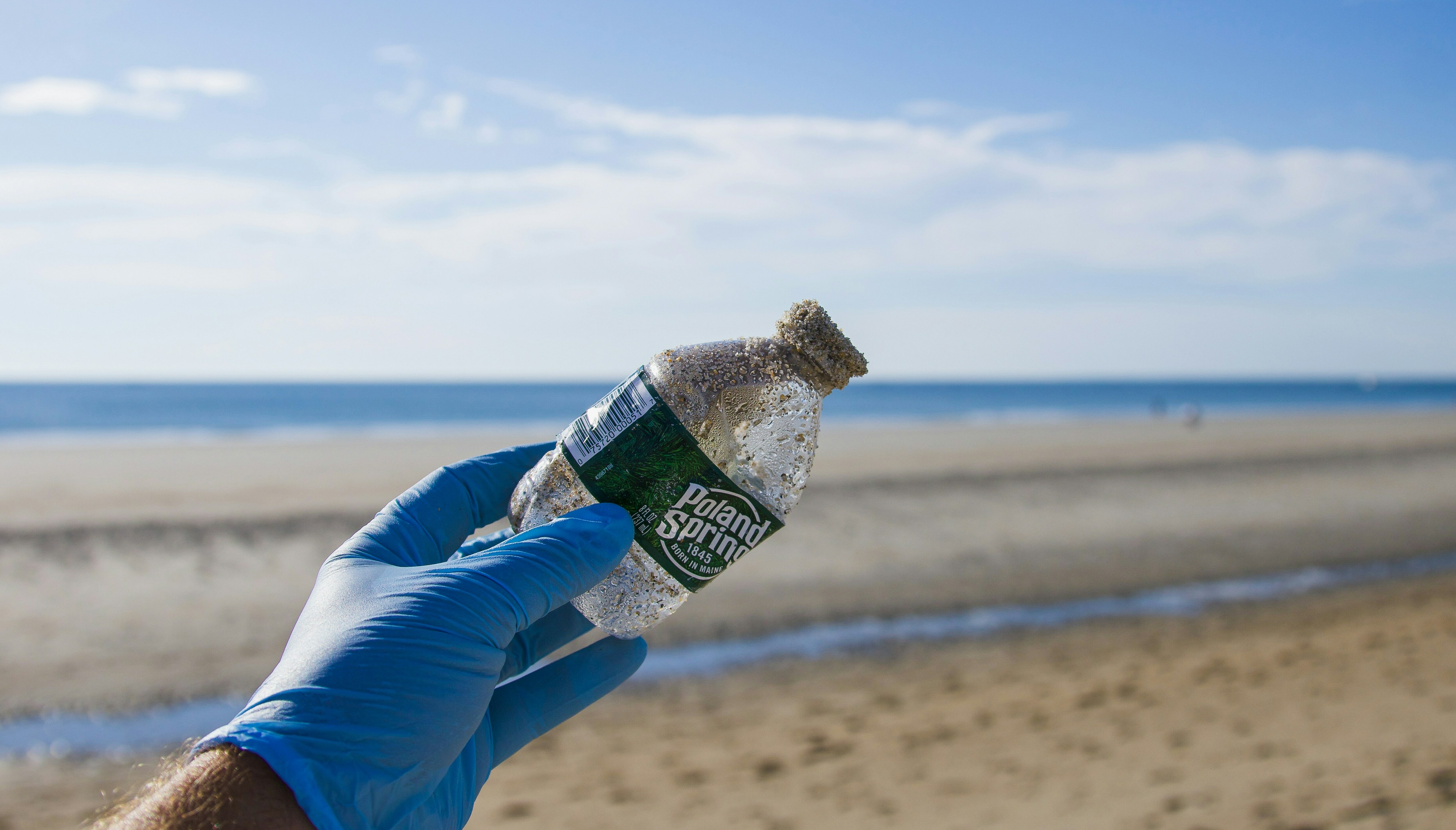 An image of a beach clean up with a hand reaching out holding a used plastic bottle. 