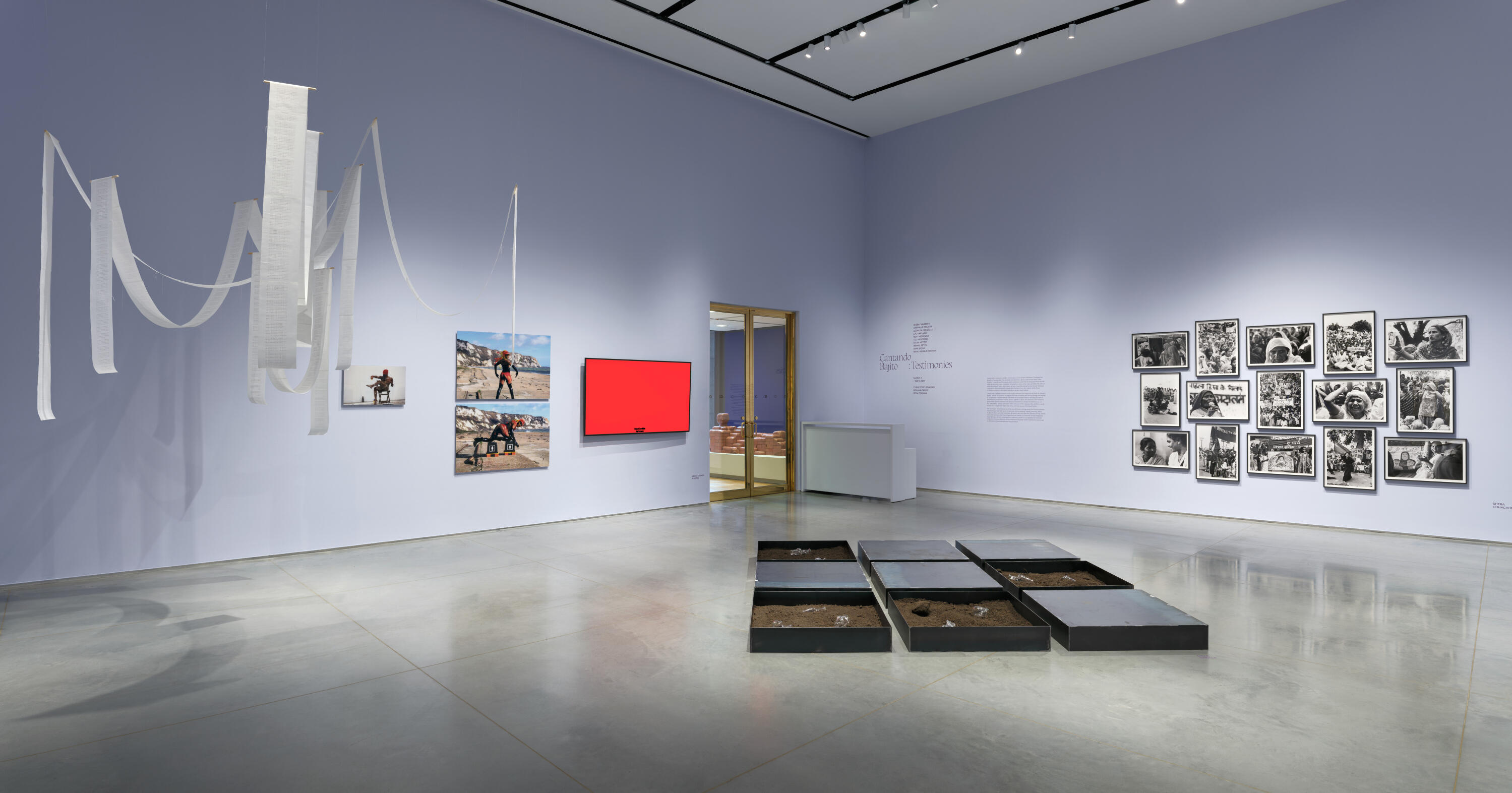 Gallery interior with light lavender walls and a gray floor. There are several artworks hung on the wall such as three printed photographs, a red monitor, and an arrangement of fifteen framed black-and-white photographs. A three by three steel-tray installation sits in the middle of the floor and a long-ribbon papered installation hangs to the left corner of the photo. In the background above a white desk, the wall has text with header “Cantando Bajito: Testimonies”. 