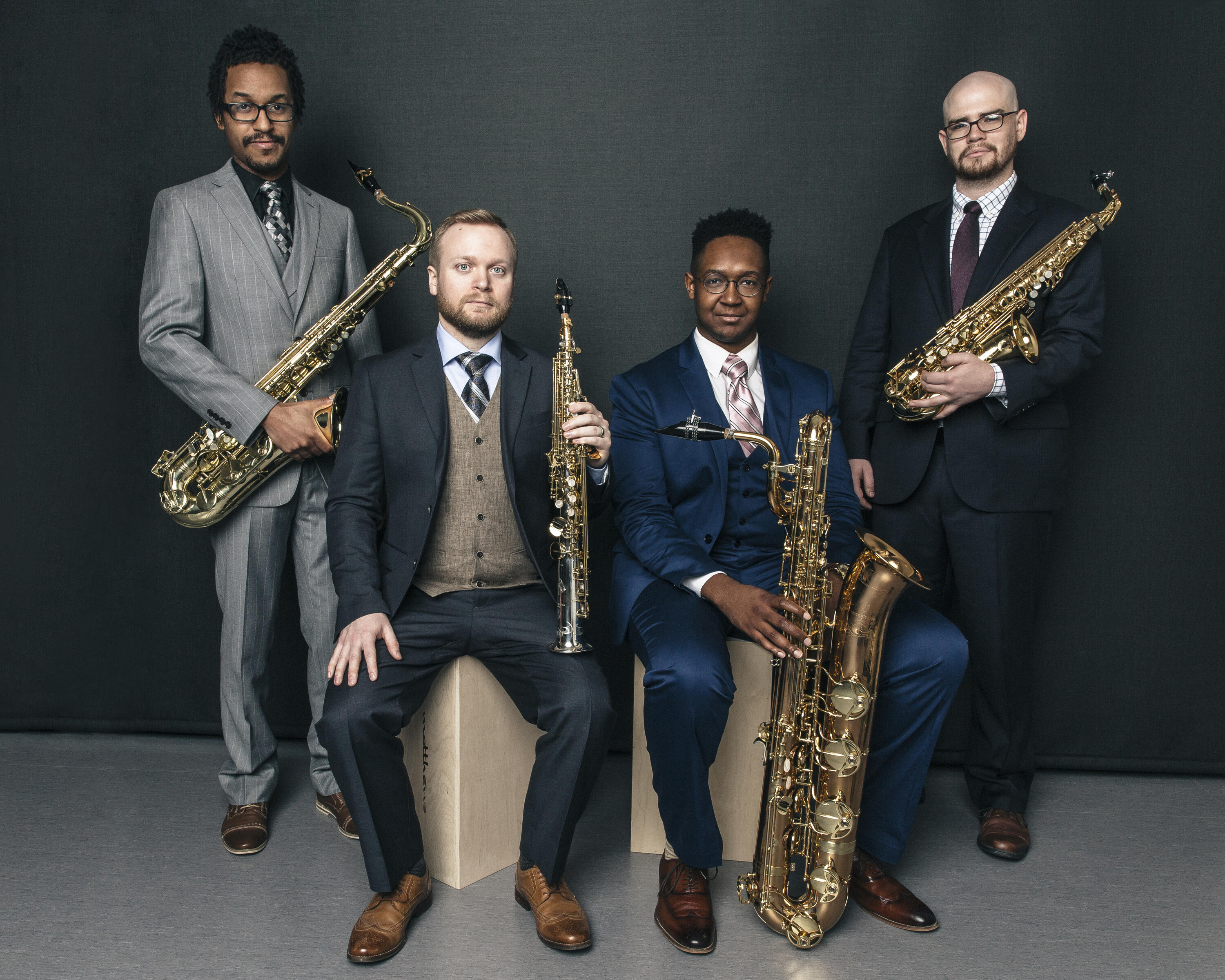 In front of a dark grey background, wearing chic suits, two members of the Kenari Quartet stand flanking two seated members, all hold their saxophones.