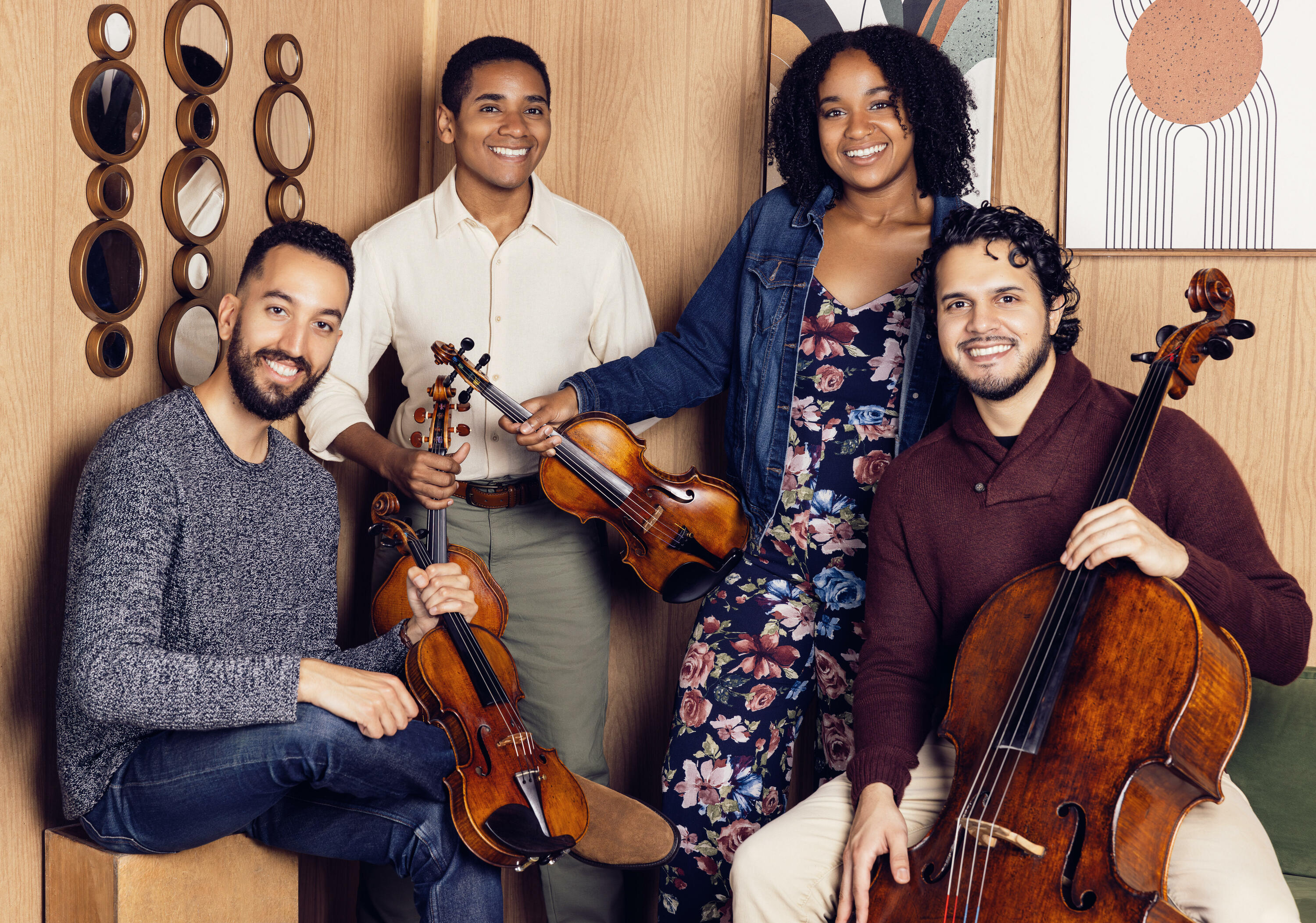 Casually dressed in colorful clothing in light wood-panelled room, the members of the Ivalas Quartet (L. to R. MENA man, black man, black woman, and Latino man) smile at viewer, holding the instruments of a string quartet.