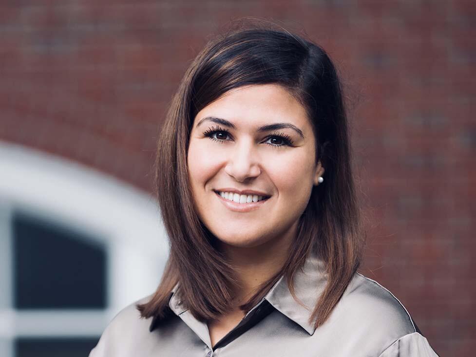 Headshot of a young brunette woman. She wears a grey blouse and there is a brick building in the distance behind her.