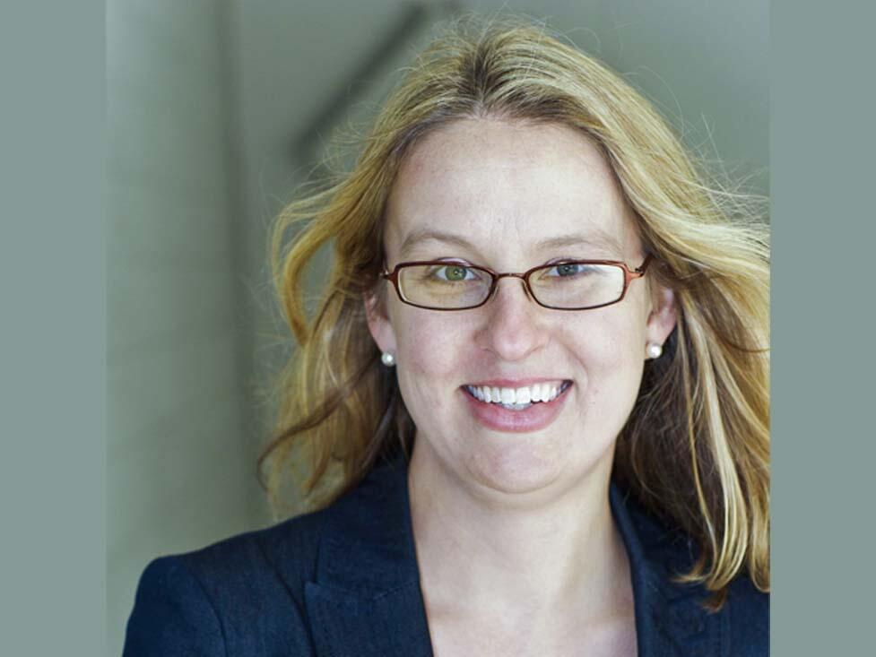 Headshot of a blonde Caucasian women in a suit with a red shirt underneath. She has long hair and wears glasses