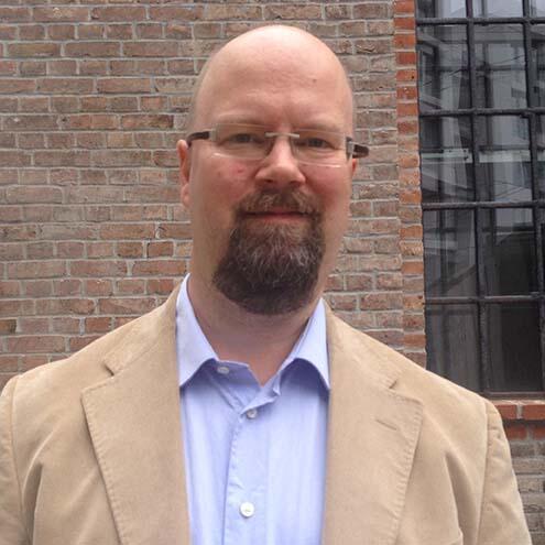 A bald Caucasian man with a goatee and glasses. He wears an open-collared dress shirt and a sport coat. He is standing in front of a brick building and a window is visible over his left shoulder.