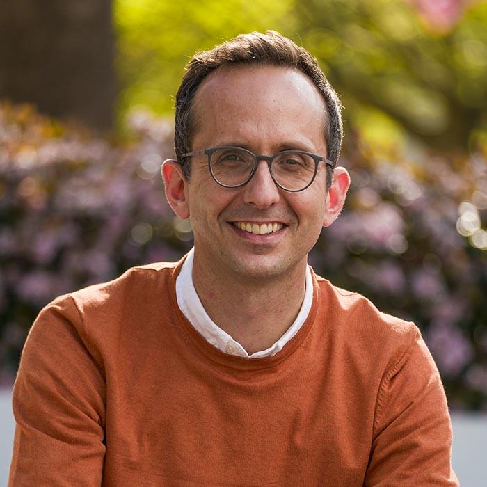 A middle-aged man with short brown hair. He wears glasses and a rust crewneck sweater with a shirt collar peeking out.