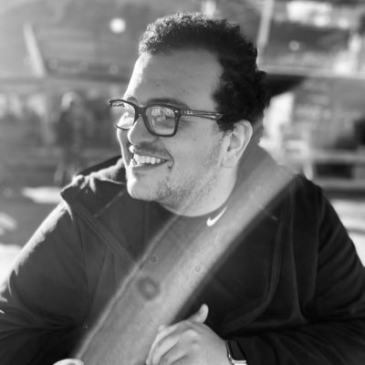 A black-and-white photo of a middle-aged Hispanic man with dark curly hair and black-framed glasses. He is outside wearing a turtleneck shirt and a jacket.