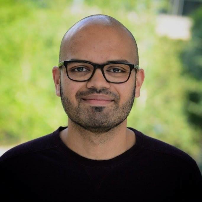 A bald man with a dark beard starting to grow in. He wears glasses and a black crewneck sweater.