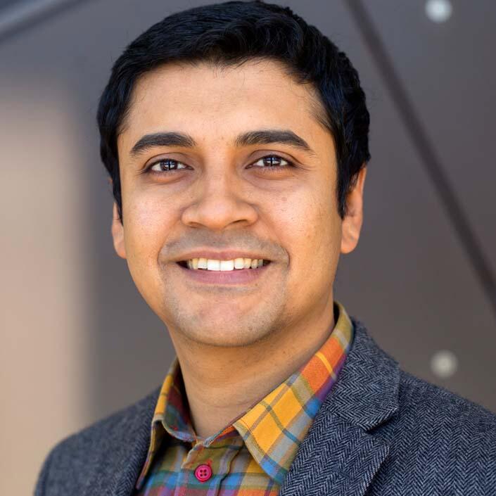 An olive-skinned middle-aged man with dark hair wears a brightly colored plaid shirt and a sport coat