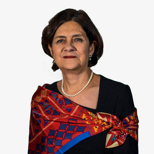 An older Hispanic woman with chin-length black hair wears a black dress and a brightly-colored scarf around her shoulders