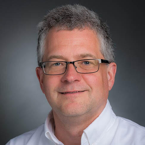 A middle-aged Caucasian with grey short hair wearing glasses and an open-collared white shirt