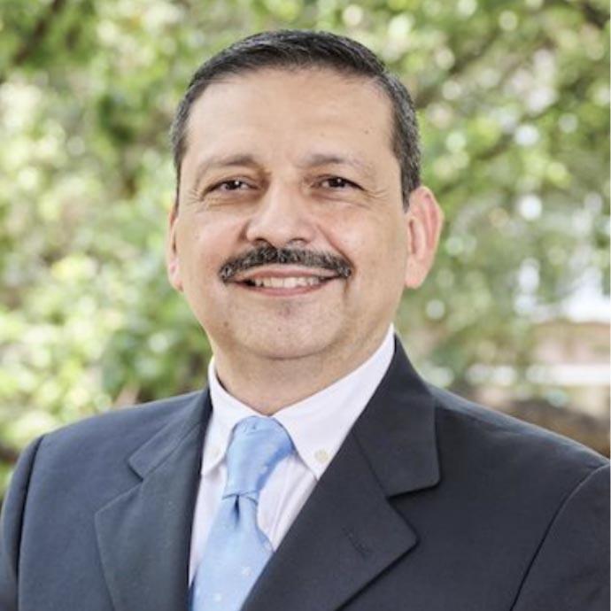 A middle-aged Hispanic man with short dark hair and a mustache, both with a touch of grey. He wears a suit and tie and is standing outside. There is a tree visible behind him.