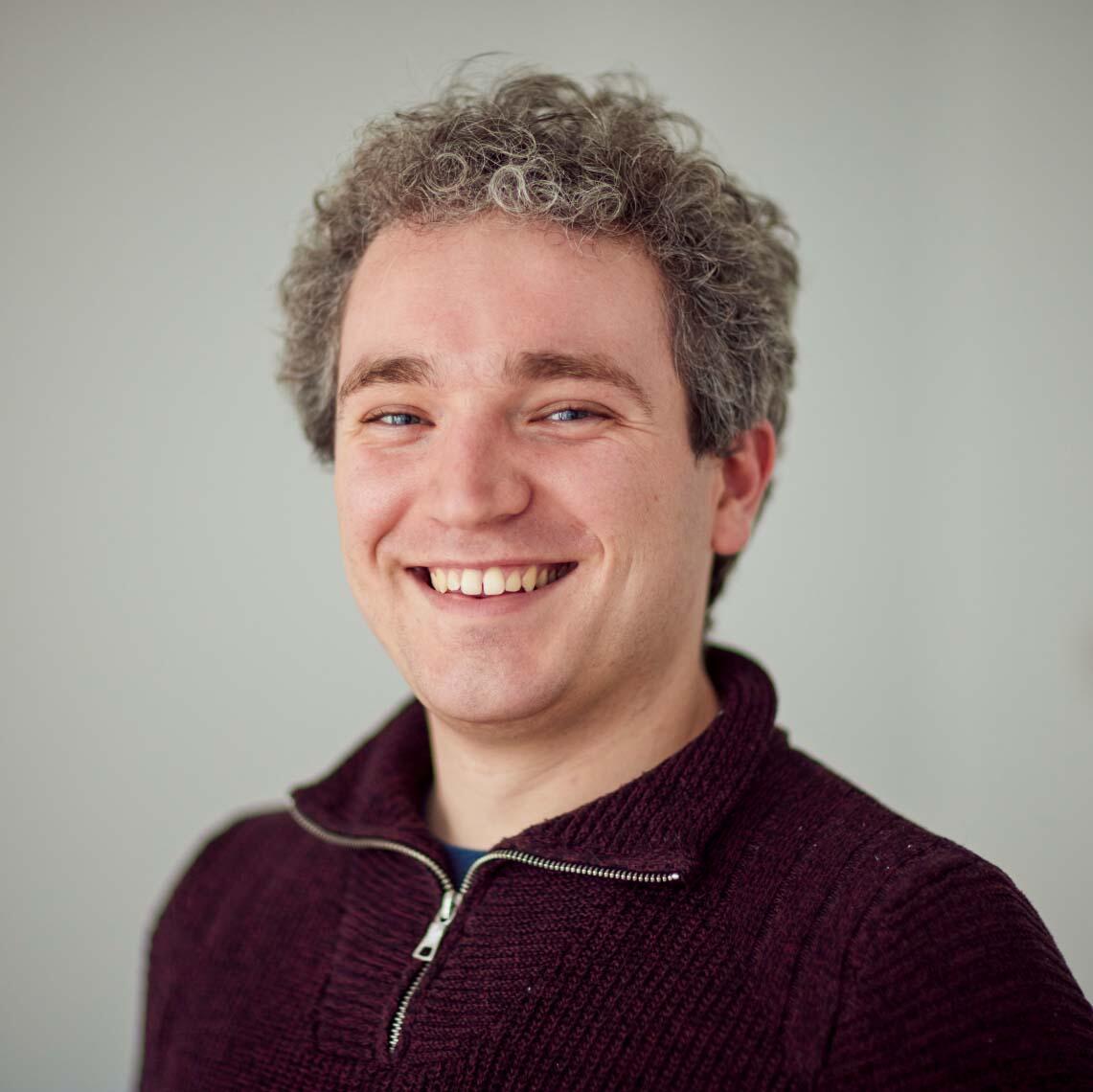 A Caucasian man with grey curly hair. He wears a burgundy zip-front sweater.