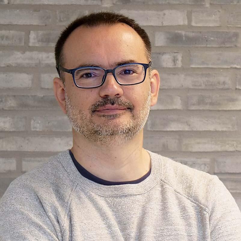 A middle-aged Caucasian man wears a casual sweater and glasses. He has dark short cropped hair and a greying beard.