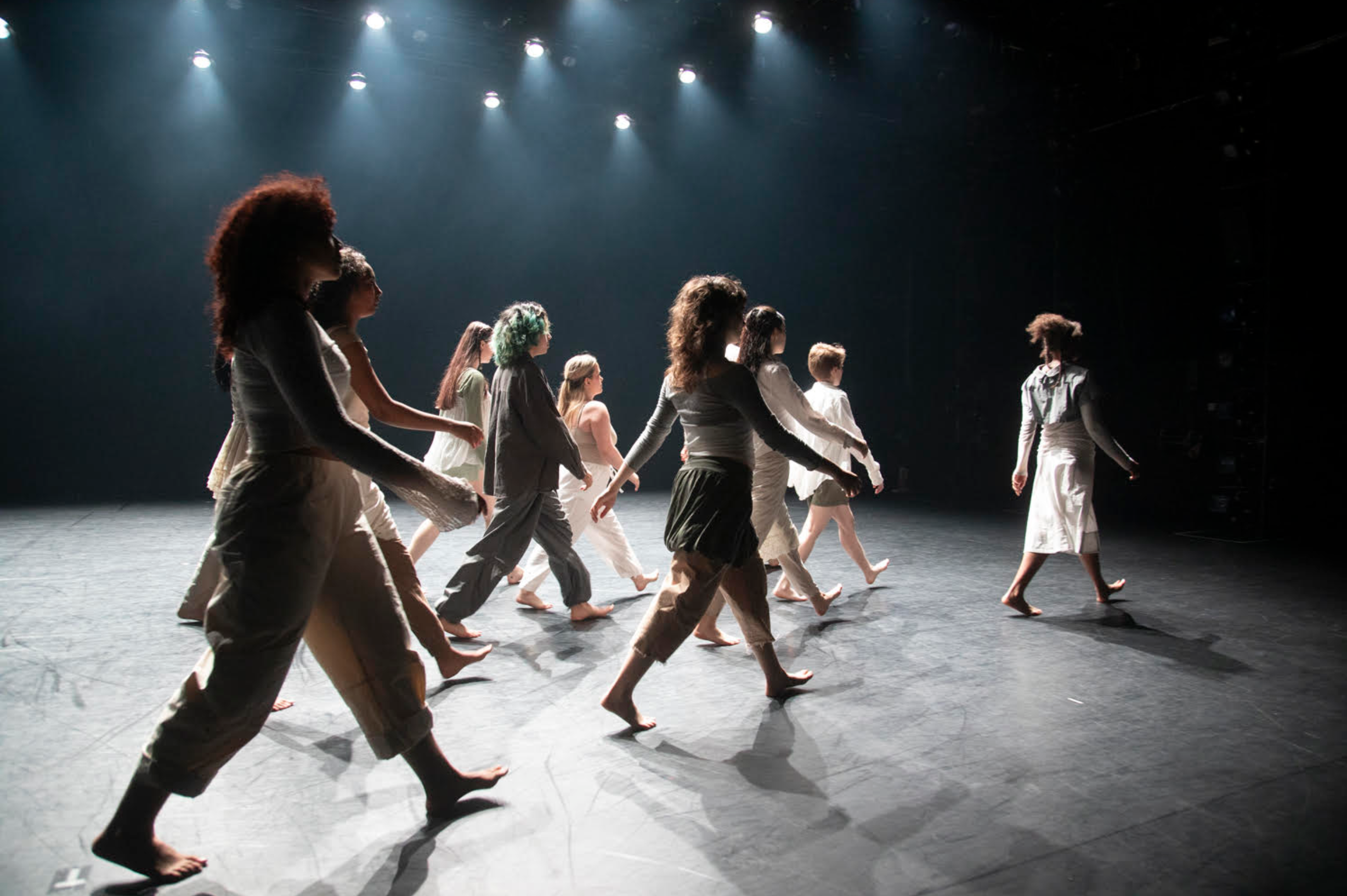 A group of four students dancing and leaping across a stage. They are wearing shiny, metallic pants and T-shirts..