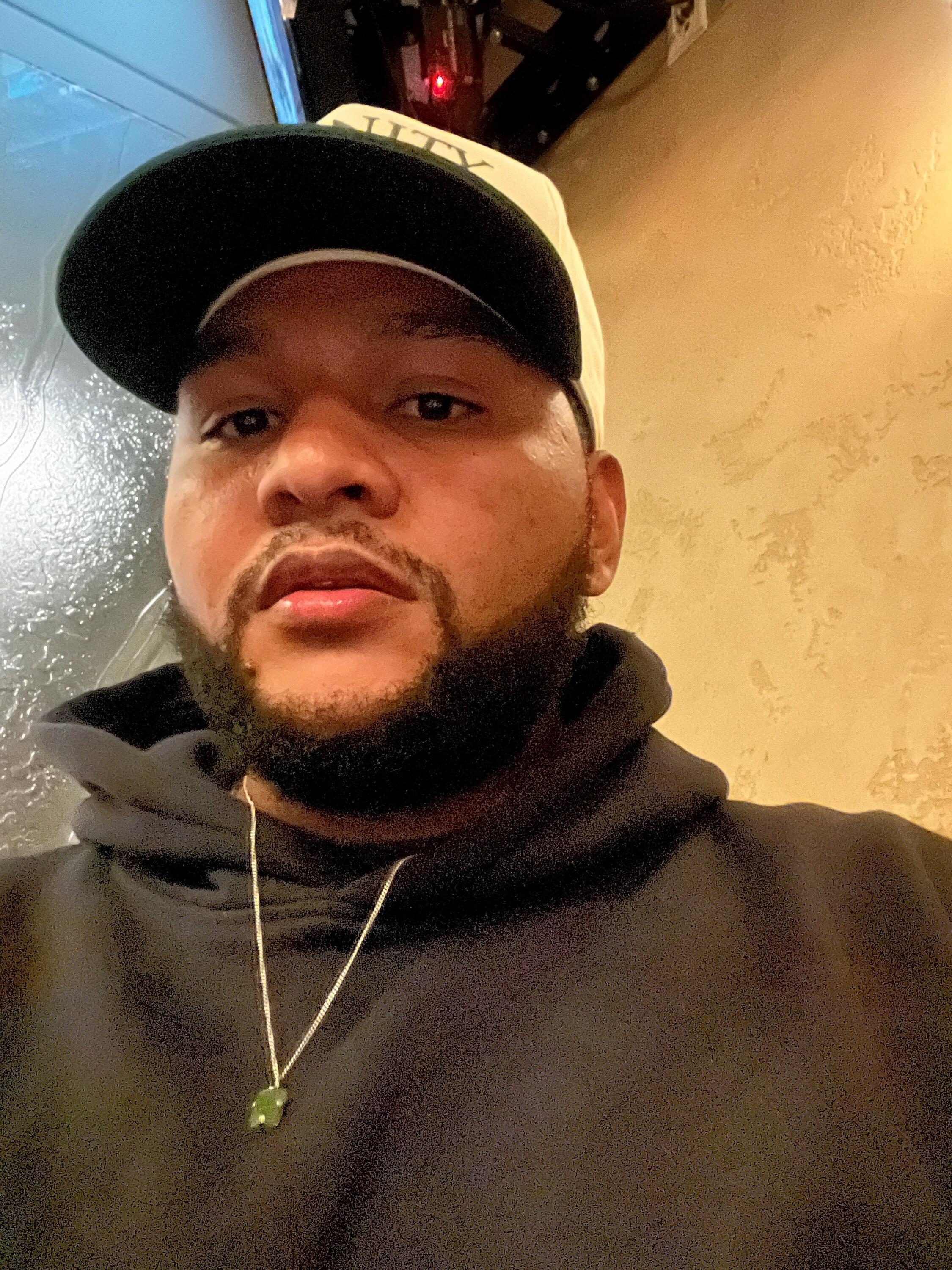 Isaac is a man with a black beard wearing a baseball cap, hooded sweatshirt, and a gold chain. 
