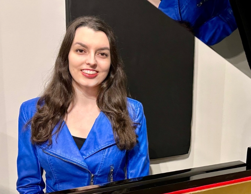Shelby is a woman with medium length brown hair wearing a blue leather jacket sitting at a piano.