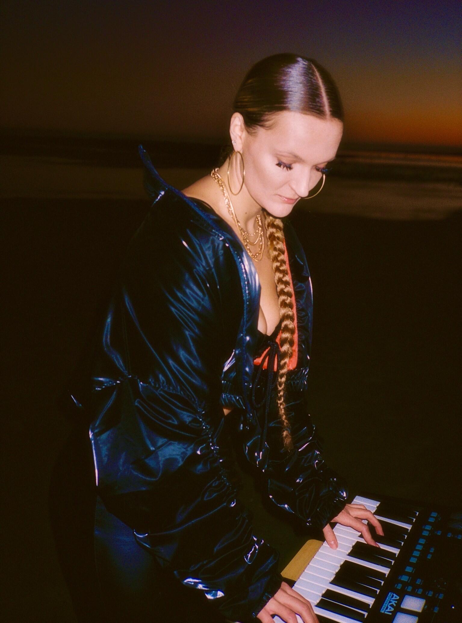 Shake is a woman with long blond hair in a braid. She is wearing a leather jacket with gold hoop earrings and gold necklaces. 