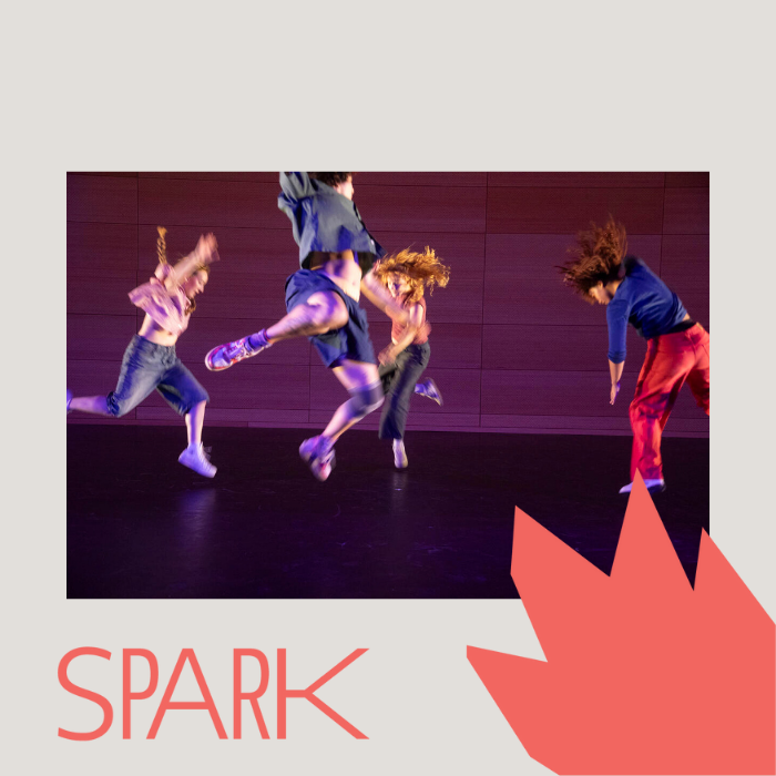 A group of four students dancing and leaping across a stage. They are wearing shiny, metallic pants and T-shirts..