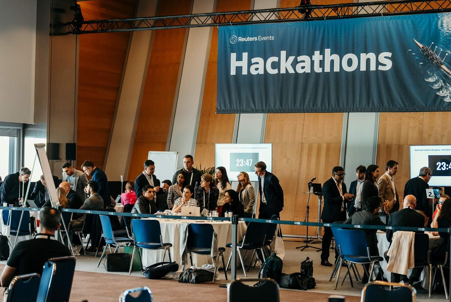 Several groups gathered around tables under a large 'Hackathons' sign