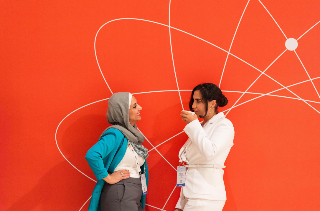 Two attendees talk in front of a red Reuters NEXT Gulf event backdrop.