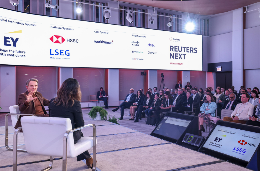 Panel discussion at Reuters NEXT, with audience members seated beneath a sponsor display.