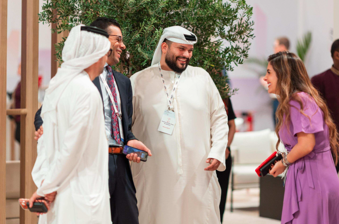 Four Reuters NEXT Gulf attendees chat near a large indoor tree, including two men in traditional Emirati dress and a woman in a purple dress.
