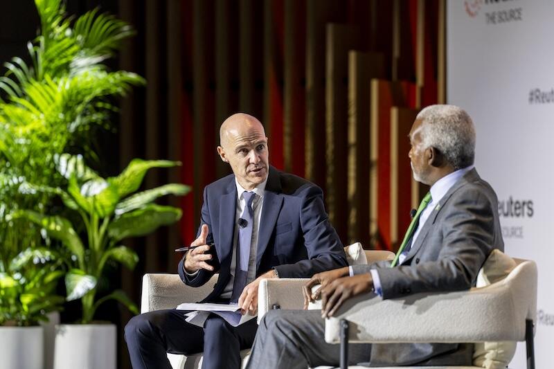 Two men in suits sit in chairs onstage having a discussion, with one gesturing while speaking; Reuters branding and plants are in the background.