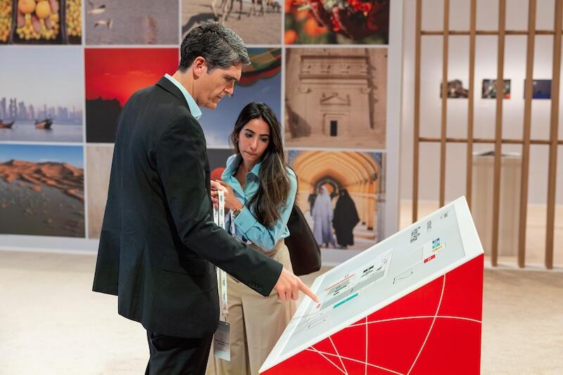 A man in a dark suit points at a large white presentation board/oversized check while a woman beside him holds an admission pass ; "REUTERS NEXT" branding and photo panels are behind them.