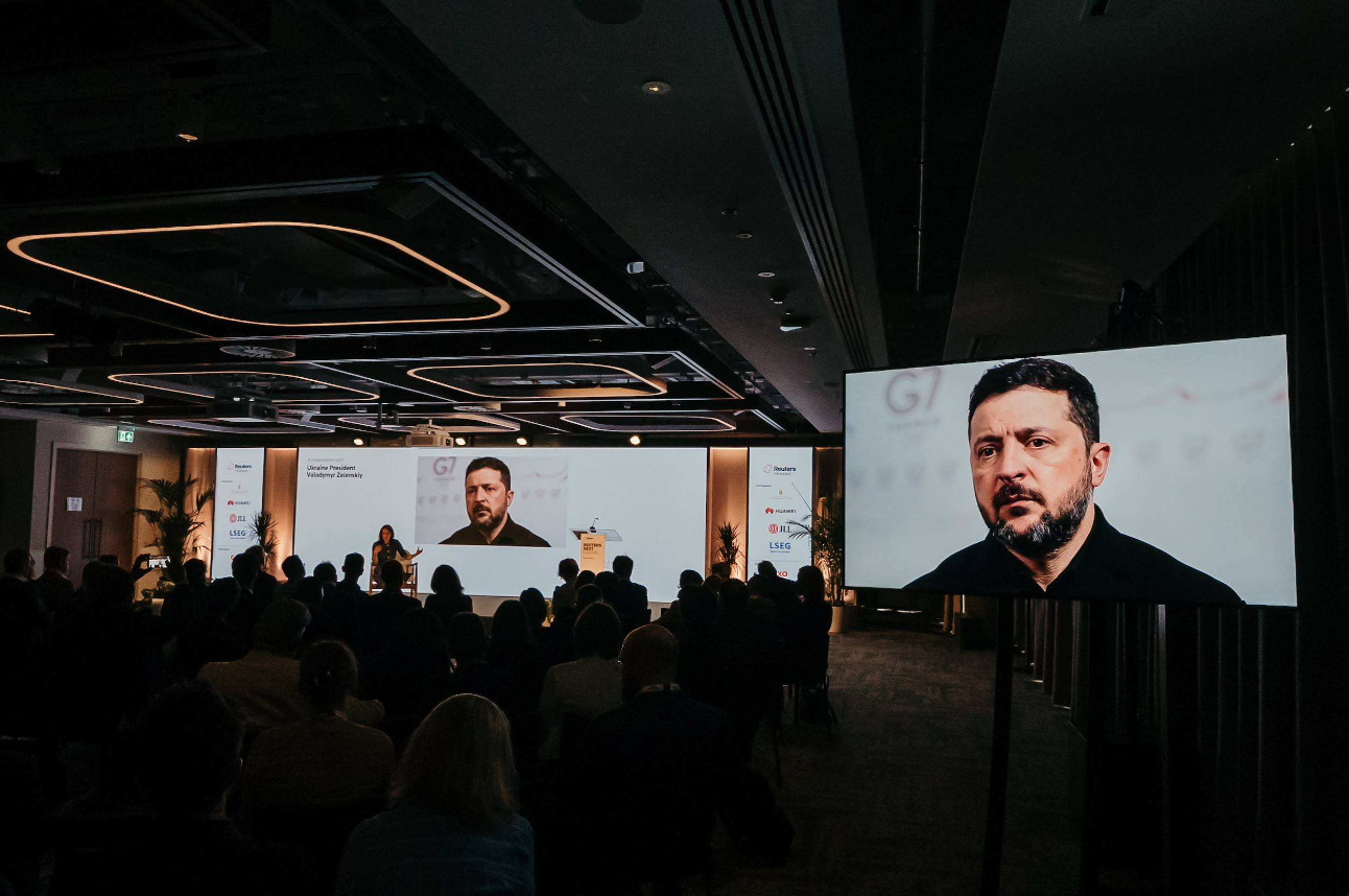 Reuters NEXT attendees gathered in a modern auditorium as President Volodymyr Zelensky of Ukraine prepares to appear via video link on dual large screens.