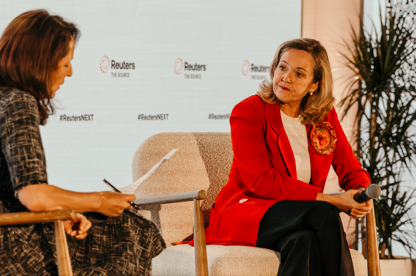 A speaker in a red blazer holds a microphone during an onstage Reuters NEXT interview, facing a moderator holding notes.