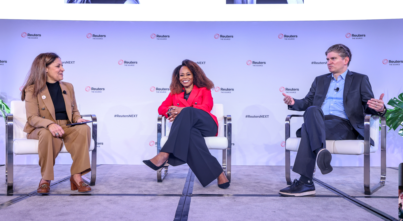 Two Reuters NEXT speakers sit onstage in conversation while a moderator in a bright red blazer listens beside them.