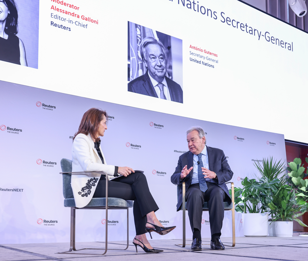 A wide shot of an audience facing a stage with a "REUTERS NEXT" screen and two people seated on stage in a moderated talk setup.