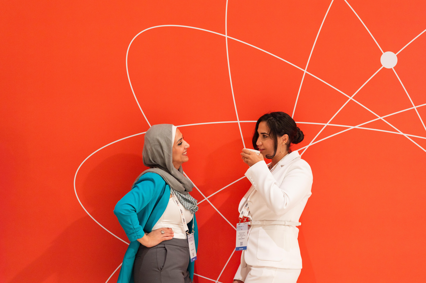 two women talking in front of a red wall with white orbit/line graphic.