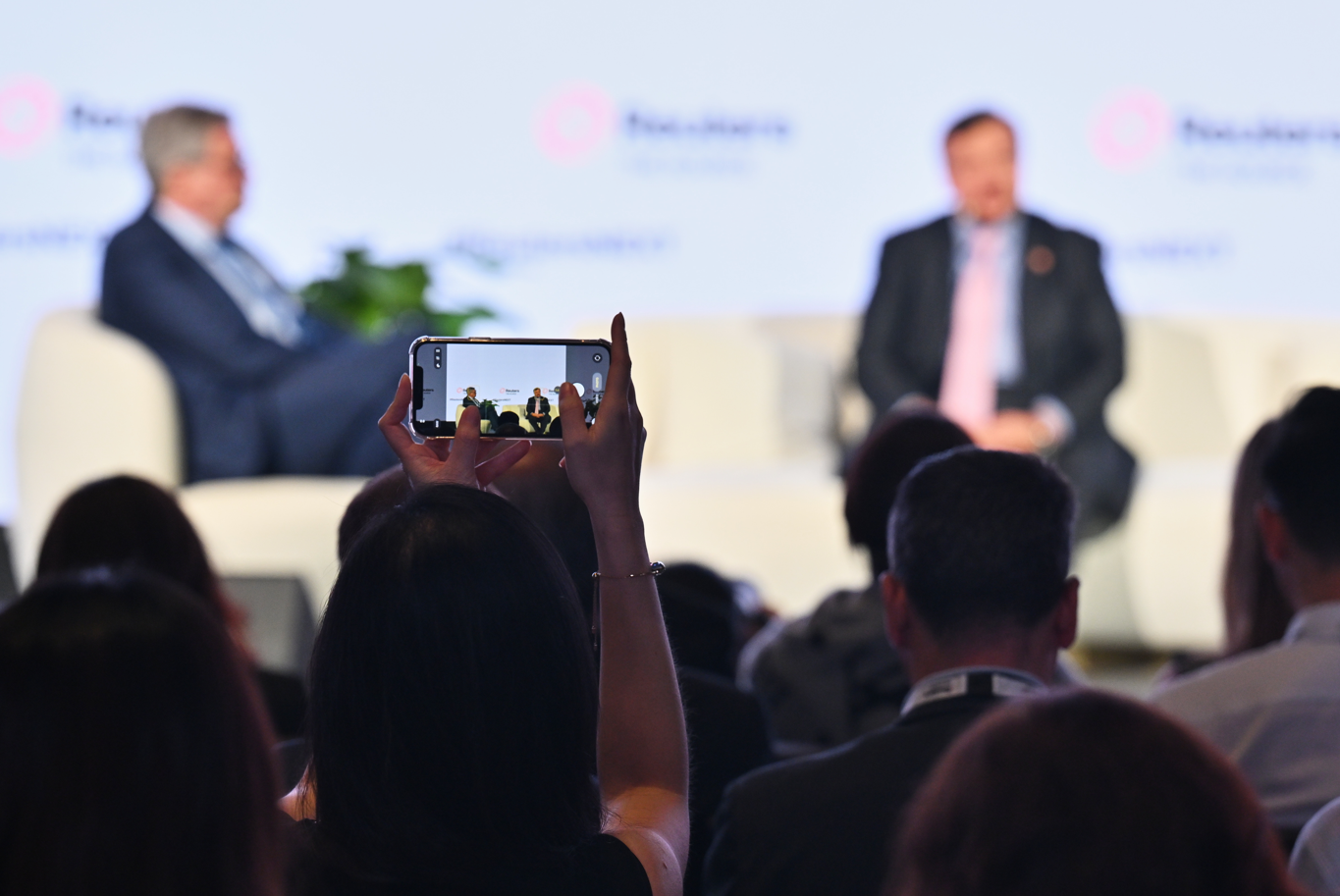 An attendee holds up a smartphone to photograph a speaker onstage at Reuters NEXT, with the speaker framed against Reuters branding and greenery.