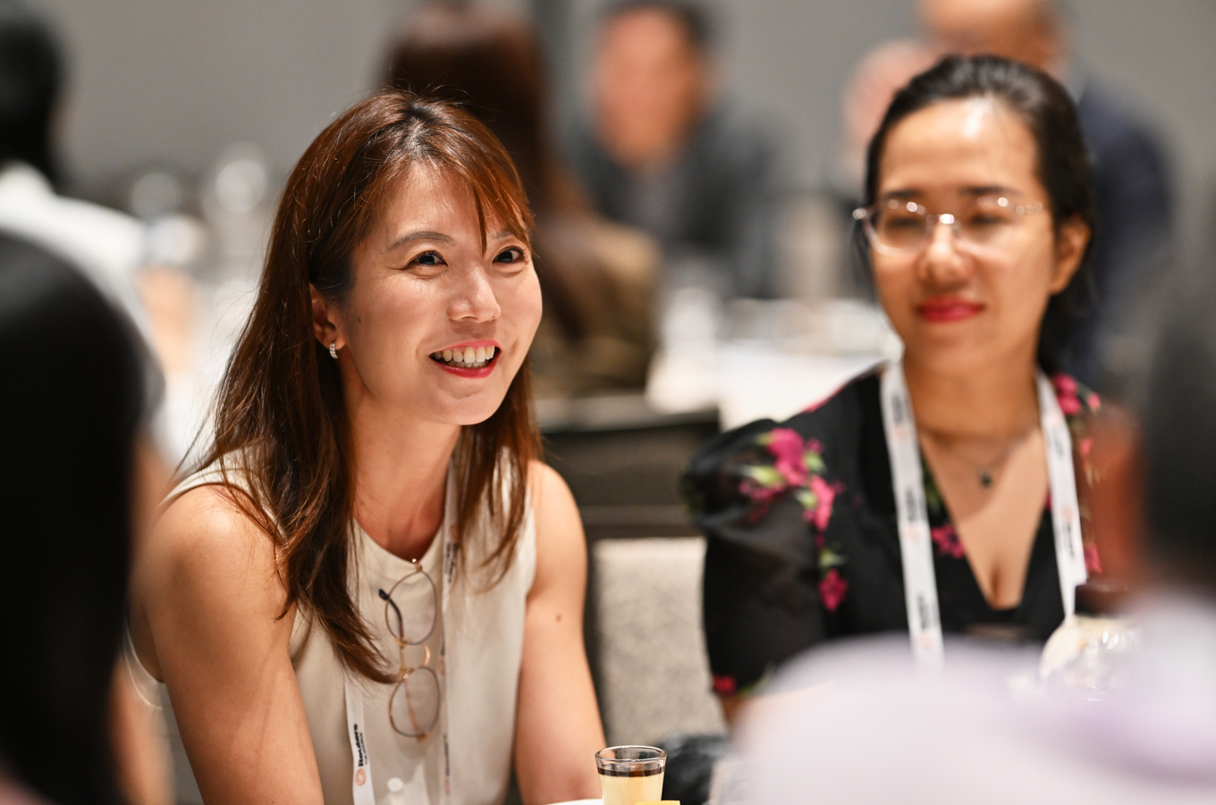 A smiling attendee in the foreground listens during a Reuters NEXT event, with other audience members softly out of focus behind her.