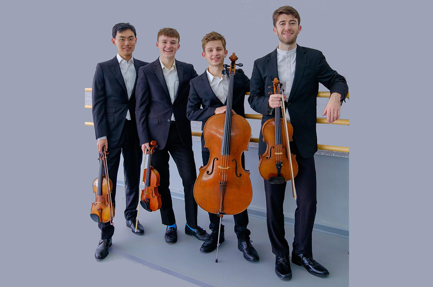 Wearing black and gazing diretly at the viewere, the members the of the Verona Quartet (an Asian man, an Asian woman, a white man and a white woman) stand outside with leafy green trees in the background, holding the instruments of a string quartet