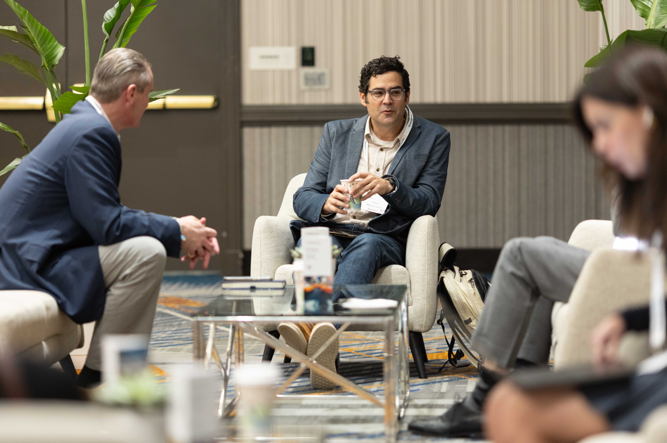 Two men seated in cream armchairs having a conversation in a hotel lounge setting. The man in the centre wears a grey-blue blazer, cream shirt, jeans, and glasses, holding a glass and wearing a conference lanyard. The man on the left wears a navy blazer and beige trousers with his hands clasped. A glass coffee table between them holds books, a cup, and small items. A patterned blue and gold rug is on the floor. A backpack rests against the chair on the right. Large tropical plants are visible in the background. A woman in grey trousers is partially visible and out of focus in the foreground right.
