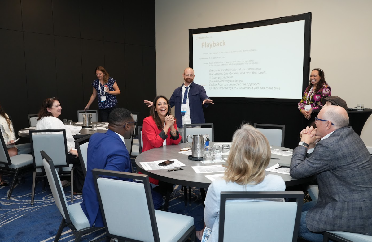 Facilitator leading a “Playback” exercise while participants at roundtables discuss and share outcomes