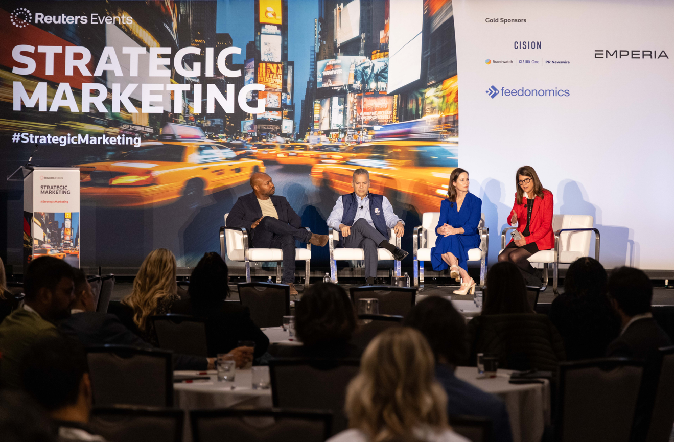 Four panellists seated in white armchairs on a stage at the Reuters Events Strategic Marketing conference. From left to right: a man in a dark navy blazer and cream top with one leg crossed; a man in a blue gilet over a light blue shirt with grey trousers; a woman in a cobalt blue dress with cream heels; and a woman in a red blazer holding a microphone, appearing to speak. Behind them is a large backdrop displaying a motion-blurred New York City street scene with yellow taxis, overlaid with the text "Reuters Events Strategic Marketing" and "#StrategicMarketing." A branded podium is visible on the far left. To the right of the stage is a sponsor board listing Gold Sponsors: Cision (including Brandwatch, Cision One, and PR Newswire), Emperia, and Feedonomics. Water bottles are placed on the stage floor between the panellists. A seated audience is visible in the foreground, with round tables and glasses of water.
