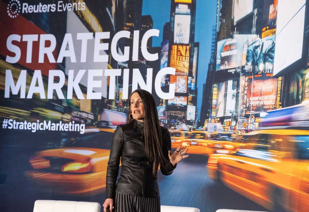 A woman with long dark hair stands speaking on stage at the Reuters Events Strategic Marketing conference. She wears a black leather jacket and a black and grey striped pleated skirt, with gold hoop earrings and a ring on her right hand. She gestures with her right hand while speaking. Behind her is a large backdrop displaying a motion-blurred Times Square night scene with yellow taxis, overlaid with the text "Reuters Events," "STRATEGIC MARKETING," and "#StrategicMarketing" in large white text. Two white armchairs are partially visible in the foreground.