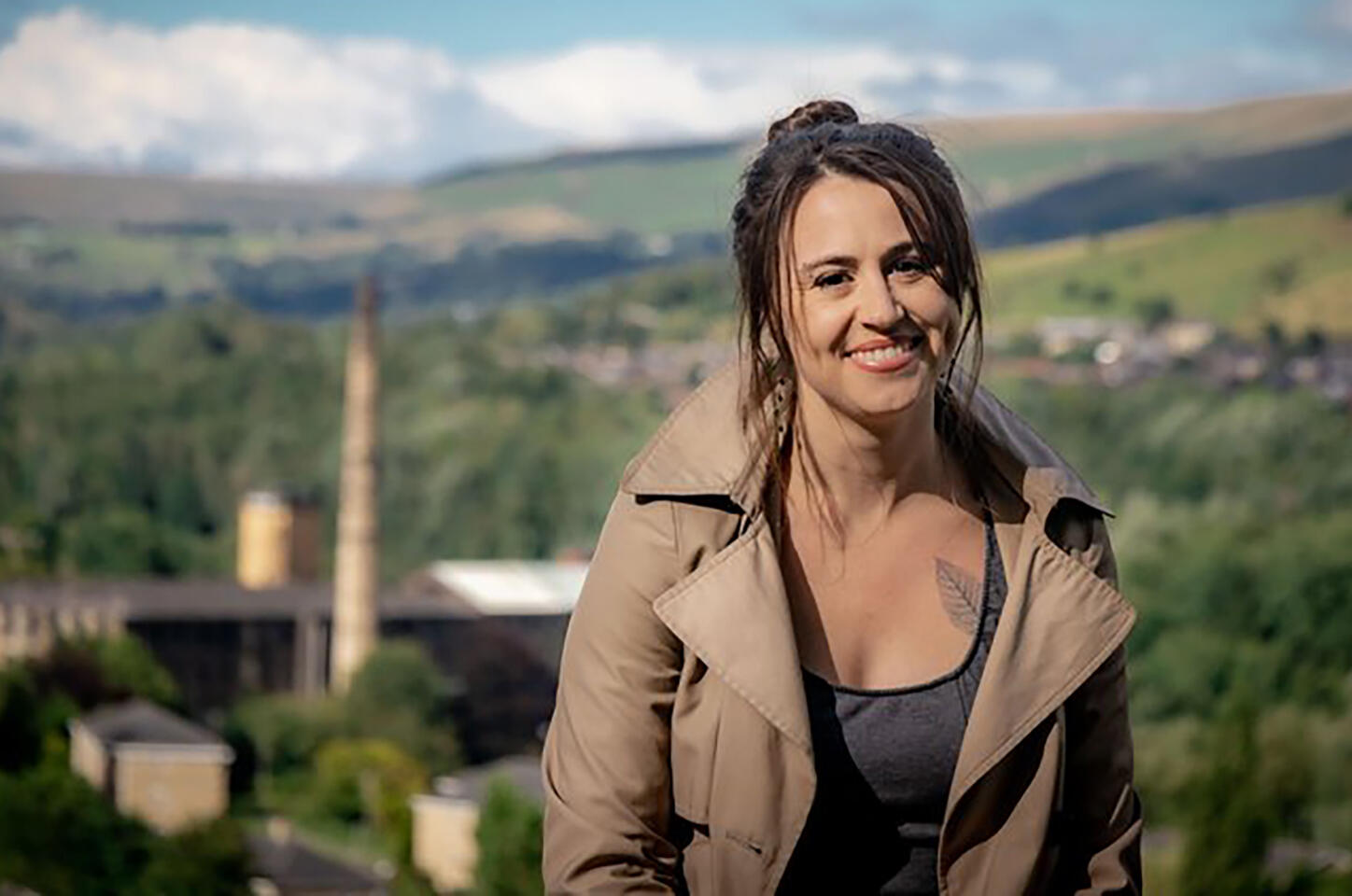 Color photo of Katherine Balch, a young white woman with pulled back long brown hair, standing on a hill with buildings in the background